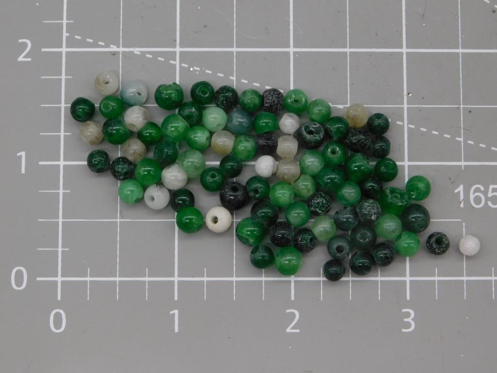Green Gemstone Round Bead Lot: Loose strand of round beads in mixed green tones (jade/aventurine-style) with some white and black accent beads, each approximately 6mm, shown on a ruled mat. Total weight labeled 18 grams. Part of