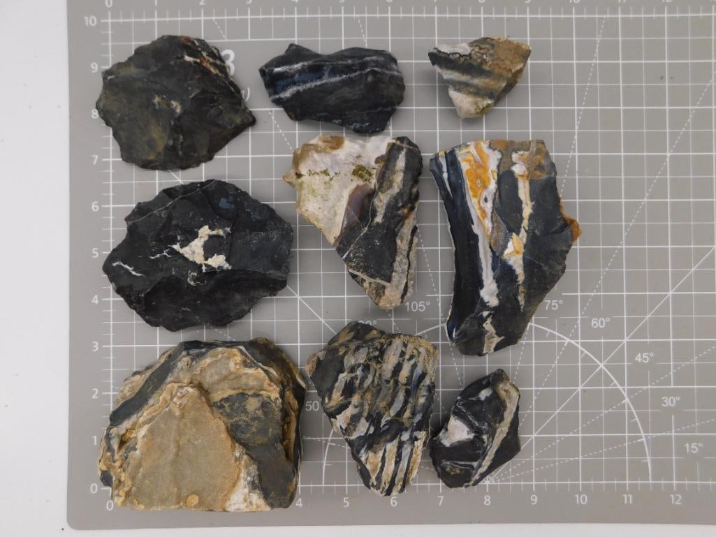 Banded Agate Rough Specimen Lot: Nine rough black and gray banded agate/jasper specimens with white and tan mineral veining, shown on a ruled cutting mat. Total weight labeled 4 lbs 4 oz 8 grams. Part of the 2026 Rock Auction.