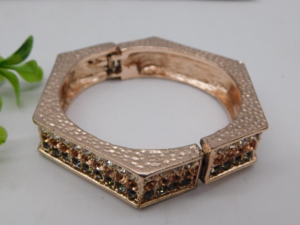 Rose Gold-Tone Hinged Bangle Bracelet with Rhinestones: Statement hinged bangle bracelet in a rose gold-tone finish with a hammered/dotted texture on top and pave-set rhinestones in champagne, clear, and smoky gray tones along the angular sides. Hexagonal 