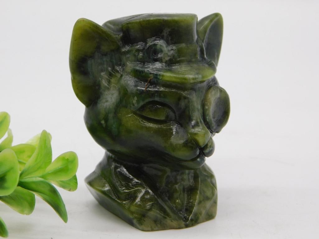 Carved Jade Figural Cat Bust in Top Hat and Monocle: A carved green jade (nephrite/serpentine) figural bust of an anthropomorphic cat wearing a top hat and monocle, with a highly polished finish showing natural mottled dark green and olive banding.