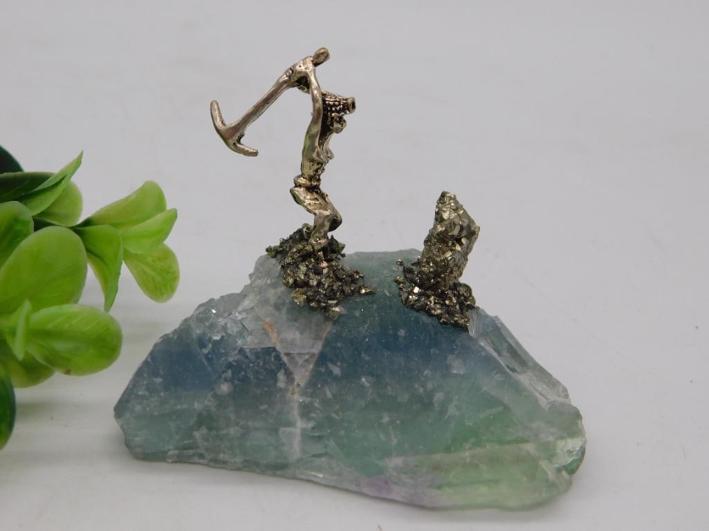 Rock Specimen with Pyrite Clusters and Miniature Metal Figurine on Fluorite Base: A rough-cut blue-green fluorite (or similar banded crystal) base displays two natural pyrite crystal clusters and a small cast pewter/silver-tone decorative figurine resembling a stylized deer or antl
