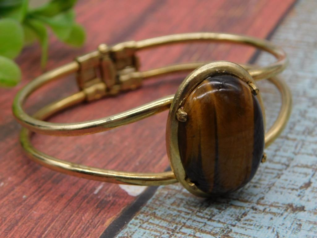 Jewelry: Gold-Tone Hinged Bangle Bracelet with Tiger's Eye Cabochon: A gold-tone metal hinged cuff bracelet set with a large oval tiger's eye cabochon in a prong setting, featuring a double-wire band design with a decorative box-link clasp. A 1966 U.S. quarter placed