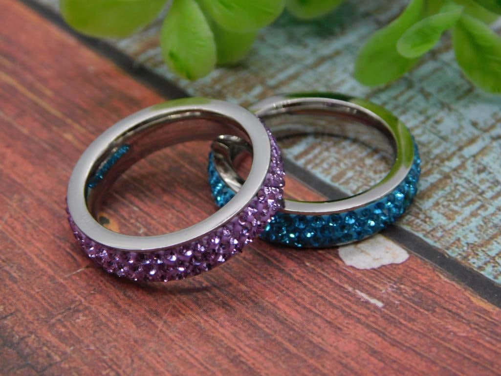 Crystal Rhinestone Stacking Rings, Set of Two: Two silver-tone stainless steel eternity band rings pave-set with sparkling crystal rhinestones, one in purple/lavender and one in blue. Shown together on a rustic wood-look surface with a quarter and