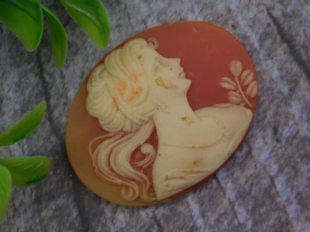 Carved Shell Cameo Brooch/Pendant - Woman's Portrait: A hand-carved shell cameo depicting a woman's profile with flowing upswept hair and a floral spray, in cream relief on a rust-orange ground. Oval shape measures roughly 1.5 inches, shown beside a