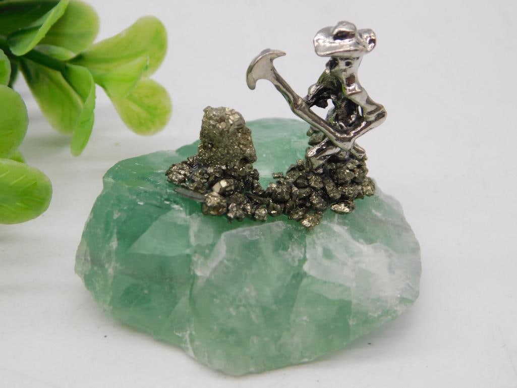 Mineral: Green Fluorite Specimen with Pyrite Cluster and Silver Miner Figurine: Rough natural green fluorite crystal specimen, roughly 3 inches across, topped with a cluster of metallic gold pyrite crystals and a small silver-tone pewter figurine of a prospector swinging a pickax