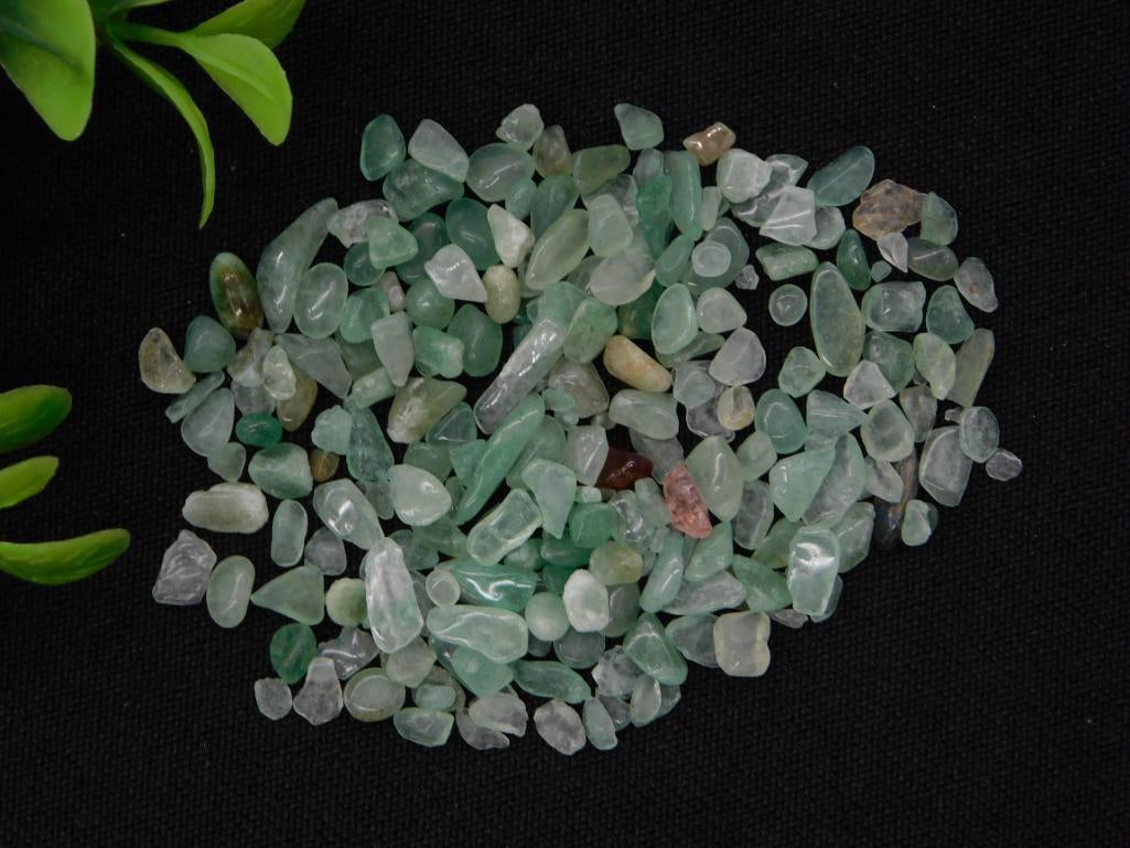 Rock and Mineral Lot - Tumbled Green Aventurine Chips, 12 Grams: A small parcel of tumbled green aventurine quartz chips and chunks with a few scattered pink and brown accent stones, displayed on black fabric. Labeled "Aventurine, 12 Grams" on the accompanying auct
