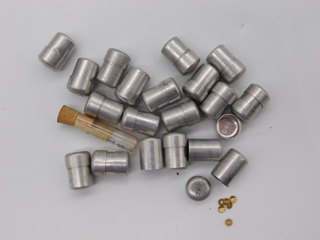 Antique - Watch JEWELS: A grouping of roughly 22 aluminum-colored cylindrical collet bushings alongside a small corked glass vial and tiny brass washers. Pieces show light surface wear.