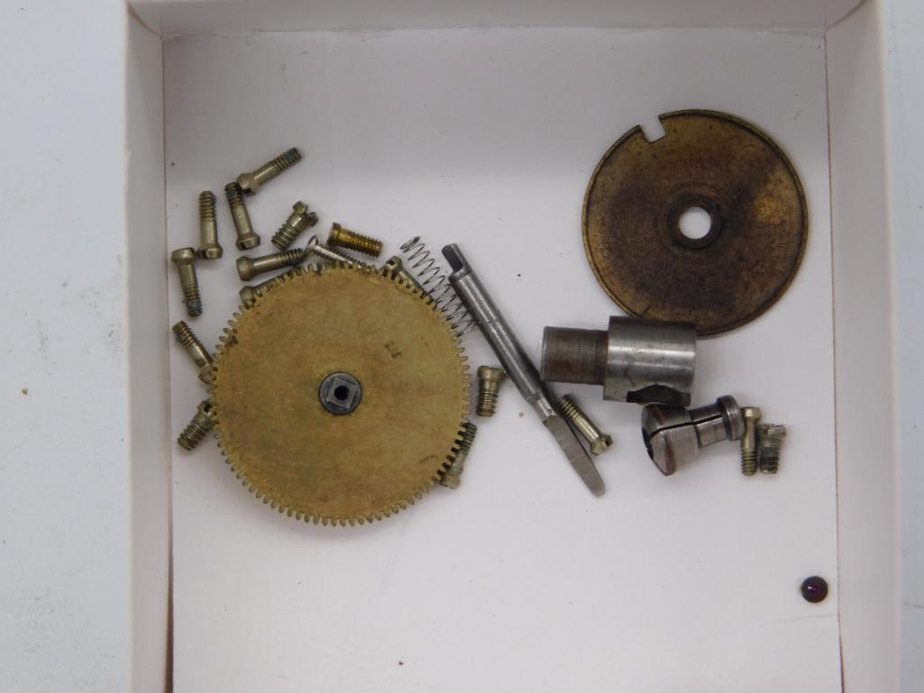Antique Clock Movement Gear and Wheel Parts Lot: A cardboard box containing a large brass clock gear wheel, a round brass plate, several threaded screws, a small spring, and assorted steel fittings. A quarter is shown for scale. The brass