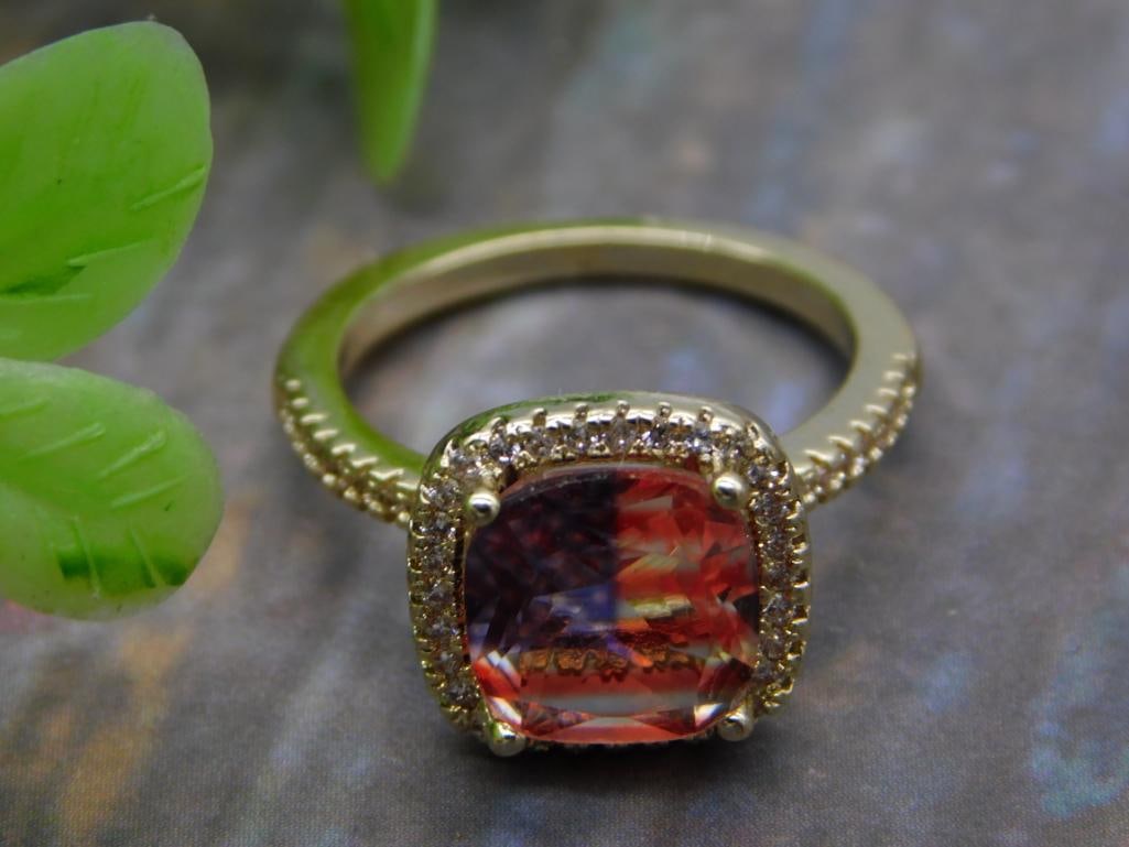 Gold-Tone Cocktail Ring with Large Orange Gemstone: A gold-tone cocktail ring featuring a large cushion-cut orange/red gemstone in a halo of small pave accent stones. Marked size 6.