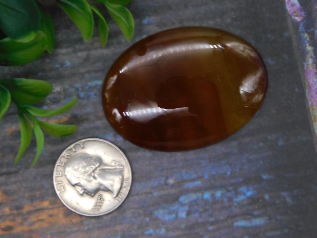 Polished Carnelian Stone Cabochon: A smooth polished oval stone in warm amber and brown tones consistent with carnelian or banded agate. Would work well as a display piece or in jewelry making.