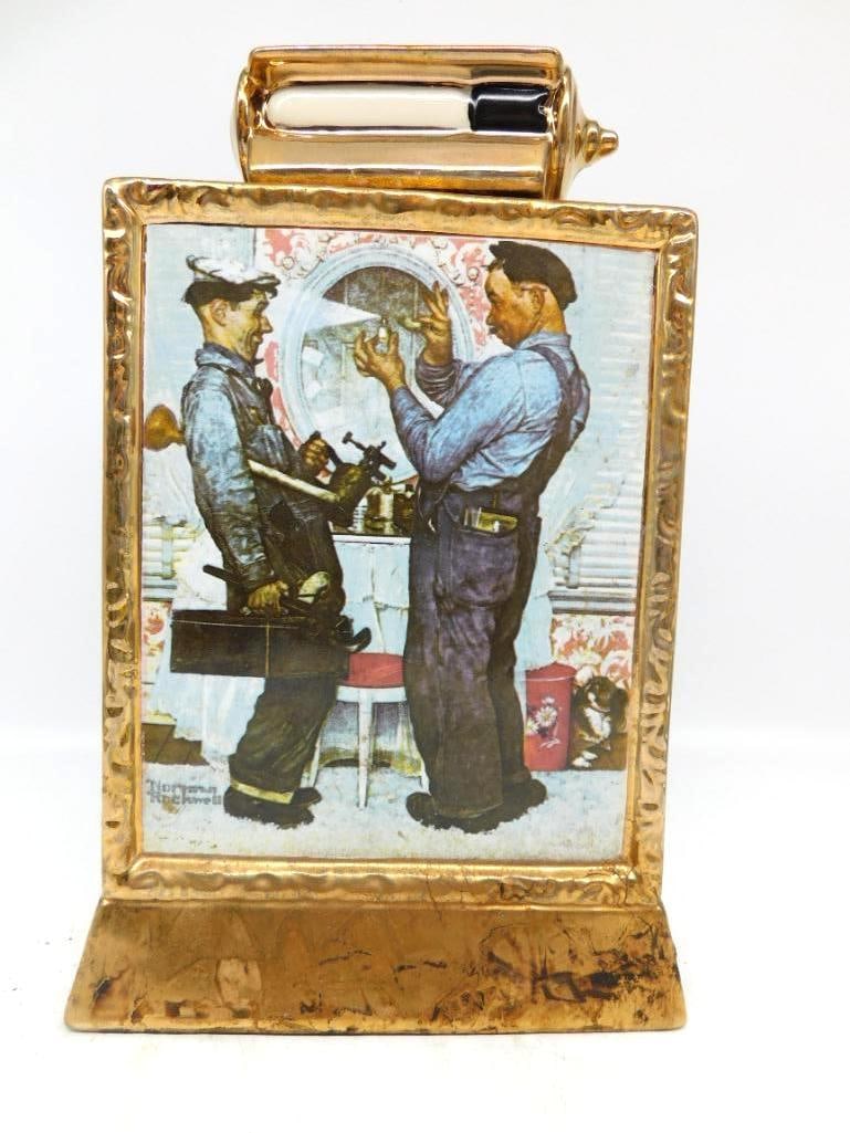 Norman Rockwell The Plumbers Ltd Ed: A ceramic decanter framed in 23K gold and bearing Norman Rockwell's The Plumbers, from the Saturday Evening Post of 1951, the two workmen pausing over a small bottle in a lady's boudoir. The gilt fram