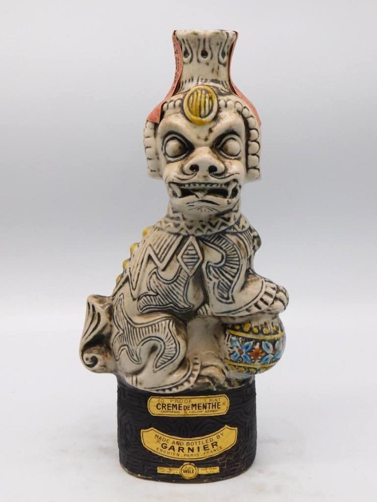 Garnier Creme de Menthe Crouching Idol: A singular Garnier decanter modeled as a crouching idol with plumed crown, bulging eyes and a fanged grin, an ornamented urn tucked beneath one arm. The incised robes are picked out in dark glaze