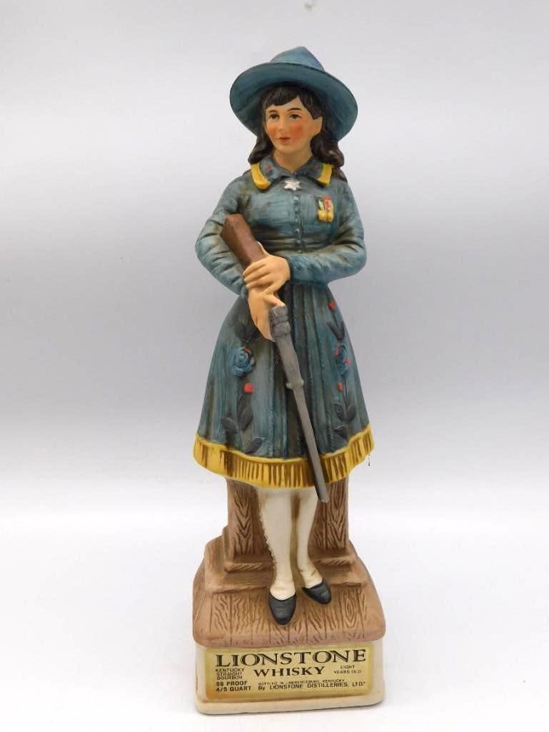 Lionstone Annie Oakley Sharpshooter: A Lionstone Distilleries decanter sculpted as Annie Oakley in period dress, rifle in hand, standing upon a wood-grain pedestal. The modeler gave her a steady, earnest bearing, and the painter followed