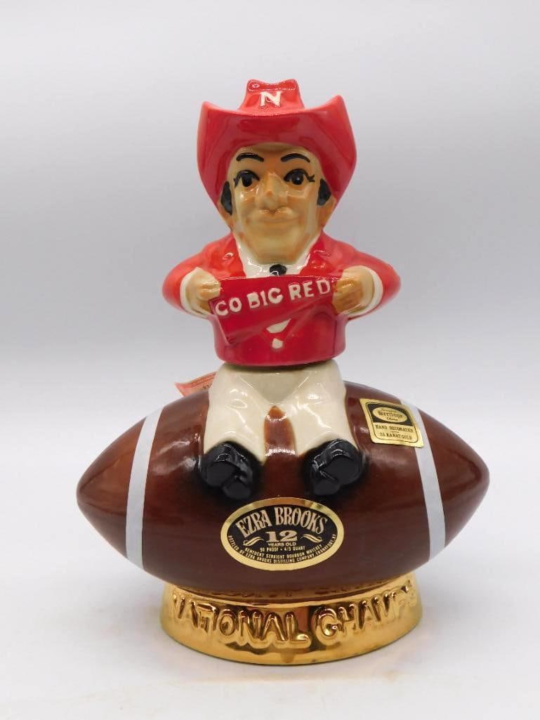 Ezra Brooks Nebraska Go Big Red Natl: An Ezra Brooks ceramic decanter shaped as a football, a Nebraska Cornhusker mascot in a red jersey perched atop it. The gold-tone base reads National Champs and Nebraska, and the bottle is marked