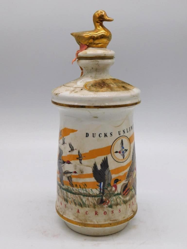 Old Cabin Still Ducks Unlimited: A porcelain Old Cabin Still decanter carrying a Ducks Unlimited wildlife scene and topped by a gold duck-form stopper. Stated honestly: some scuffing and discoloration to the body and to the neck