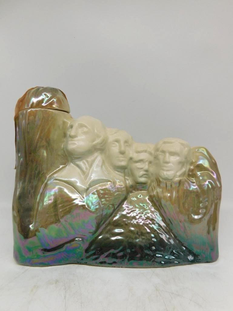 J.W. Dant Mount Rushmore Decanter in: A J.W. Dant decanter modeled as the carved face of Mount Rushmore, the four presidents in creamy white against a rock wall glazed in a shimmering mother-of-pearl luster. The reverse is embossed Mt. Ru
