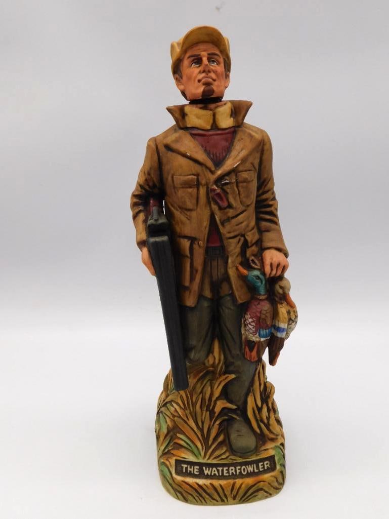 The Waterfowler Duck Hunter Decanter: A painted bisque decanter modeled as a duck hunter in his shooting jacket, shotgun in hand and a brace of ducks carried home, titled The Waterfowler on the base. Approximately 12 inches tall, with