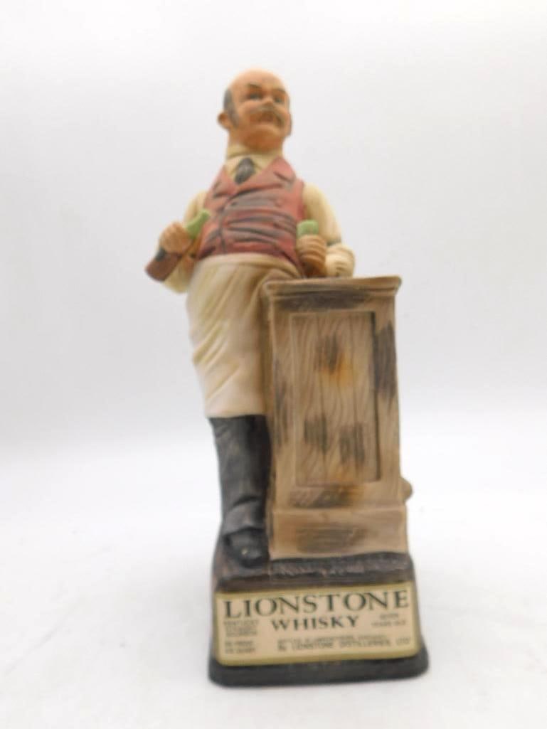 Lionstone Old West Bartender Decanter: A Lionstone decanter modeled as a frontier bartender at his plank counter, a bottle in one hand and a glass in the other, waistcoat red and apron tied behind. The sculptor gave him an obliging look,