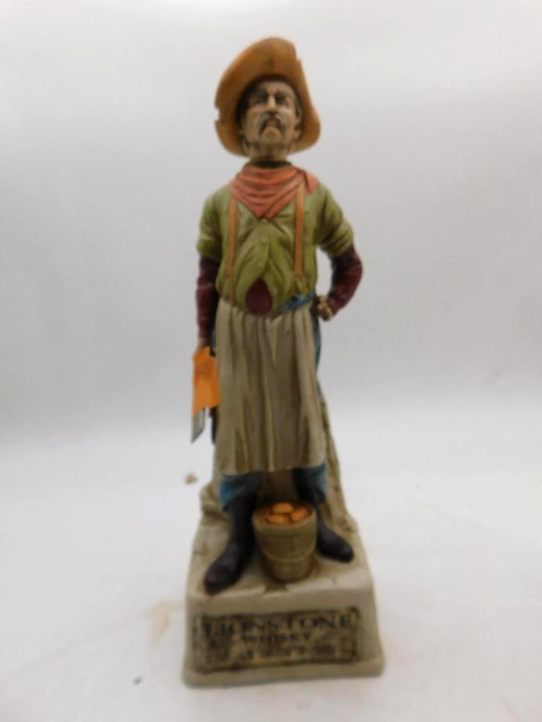 Lionstone Frontier Shopkeeper Decanter: A Lionstone decanter modeled as a frontier shopkeeper in a long apron and broad hat, cloth and cleaver in hand and a bucket of fruit set at his boots. The face is a shrewd and particular bit of