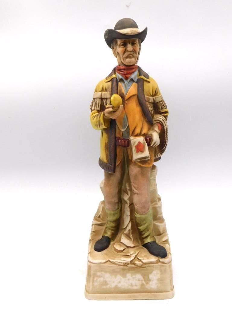Hand-Painted Cowboy Decanter with Lasso: A hand-painted ceramic decanter modeled as a cowboy in a fringed yellow jacket, a star badge at his breast and a lasso in hand, standing on a rocky base. The painter did not stint on the fringe or the