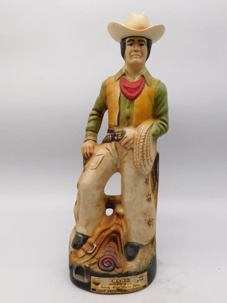 Jim Beam Cowboy in Chaps Western: A Jim Beam decanter modeled as a cowhand at rest, hat pushed back, neckerchief bright at the throat, chaps and gunbelt rendered with a shrewd eye for the working West. The muted greens and golds of th