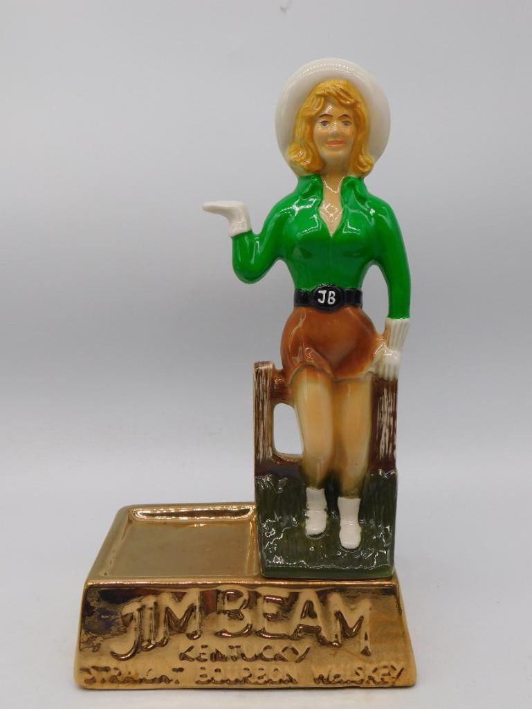 Old West Cowgirl Figural Decanter on: A hand-painted decanter modeled as a costumed Old West cowgirl in a green blouse, tan skirt and wide-brimmed hat, mounted on a wood base with a metal identification plaque. The plaque text was not