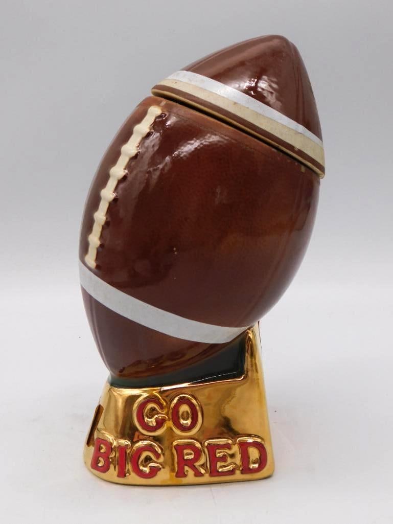 Ezra Brooks Nebraska Cornhuskers Go Big: An Ezra Brooks ceramic whiskey decanter shaped as a football lettered GO BIG RED, set upon a gold-tone trophy base marked NEBRASKA. Glossy brown and white finish, handsome enough to hold a shelf on it