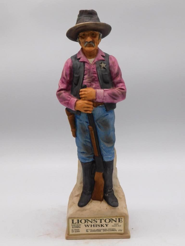 Lionstone Western Sheriff Decanter: A Lionstone Distilleries decanter cast as a Western sheriff in a pink shirt with star badge, rifle held ready. Stands approximately 12 inches tall, the Lionstone whiskey label present on the base. Lig