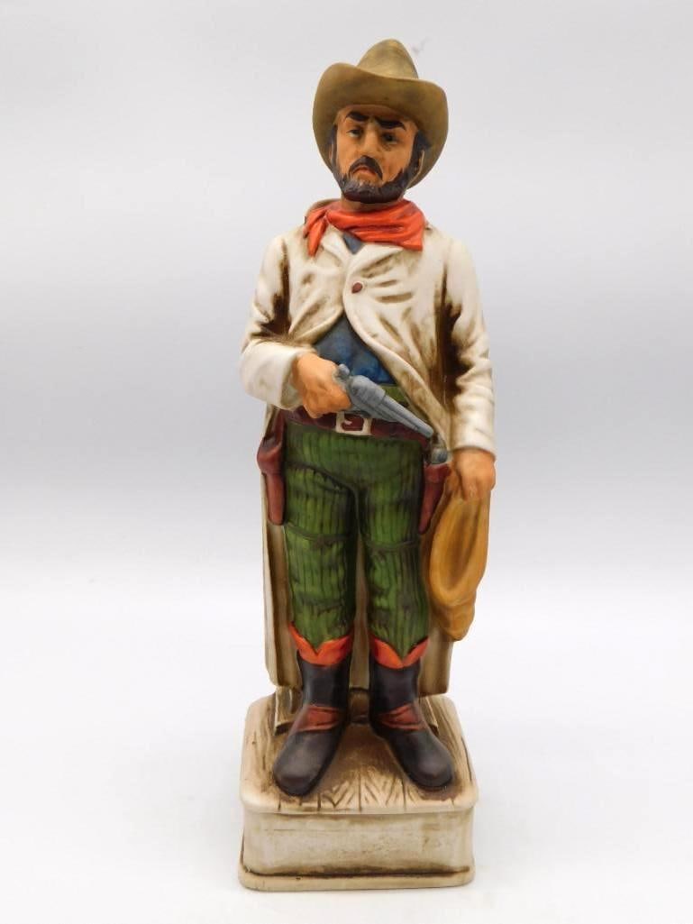 Western Gunslinger Cowboy Ceramic: A hand-painted ceramic decanter sculpted as a Western gunslinger in hat, bandana and chaps, revolver in hand, mounted upon a faux wood-grain base. An unbranded example in the manner of the 1970s