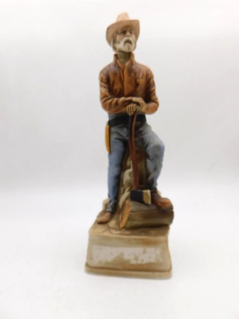 Sculpted Cowboy Decanter, Seated with: A porcelain decanter modeled as an old cowhand in fringed jacket and chaps, seated on a rock with a lariat in hand and an axe at his boot. The bisque surface is softly painted, and the sculptor gave