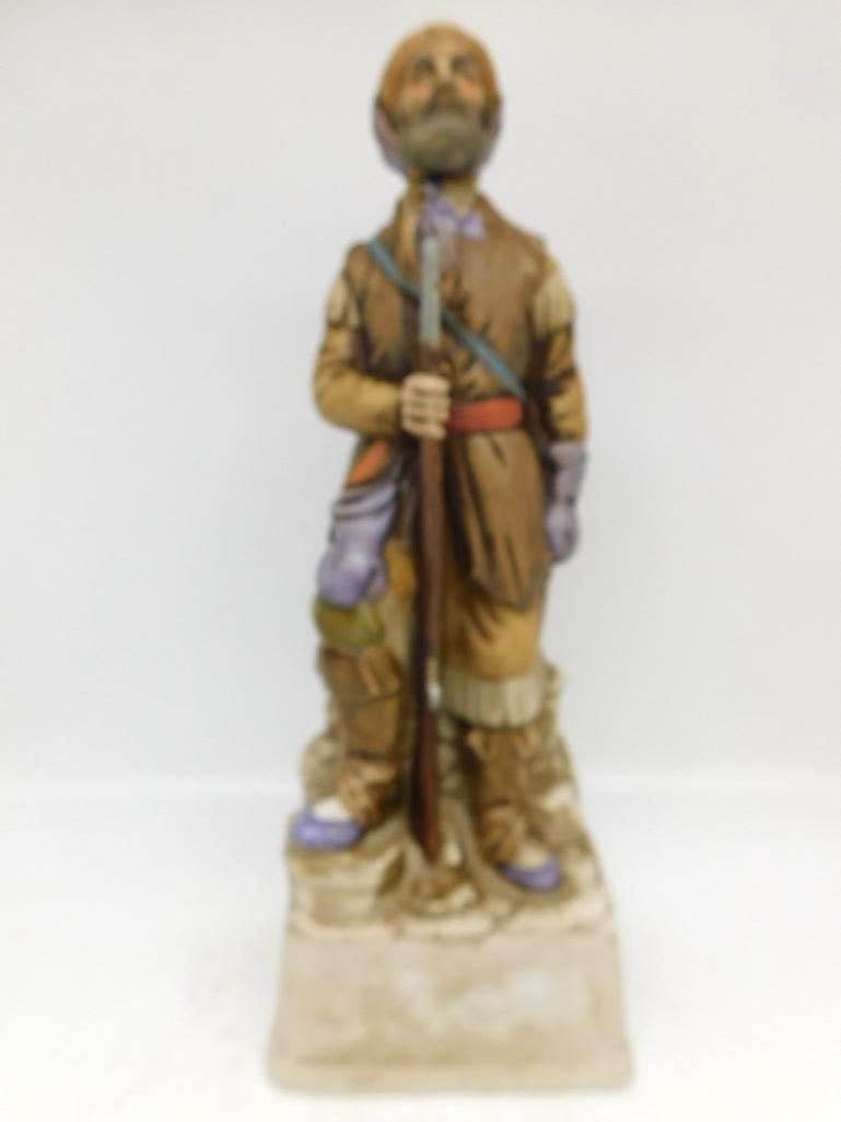 Frontiersman with Long Rifle Porcelain: A porcelain decanter modeled as a bearded frontiersman in fringed buckskin, long rifle held before him and a powder horn slung at his shoulder. The painting is soft and earthy, the tasseled leggings