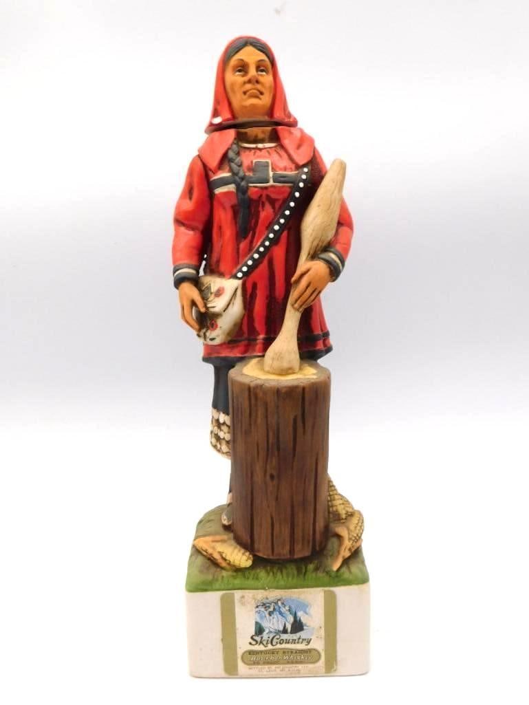 Ski Country Native American Woman: A Ski Country bourbon whiskey decanter sculpted as a Native American woman in a red hooded garment, a beaded bag at her side, standing beside a carved wood-form pedestal. The folds of the cloak are