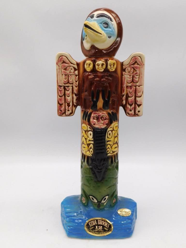 Ezra Brooks 12 Years Totem Pole Decanter: A stout Ezra Brooks decanter in the form of a Northwest Coast totem pole, crowned with a carved thunderbird head above stacked mask motifs in brown, red, and cream, all upon a blue rock-form base. Sta
