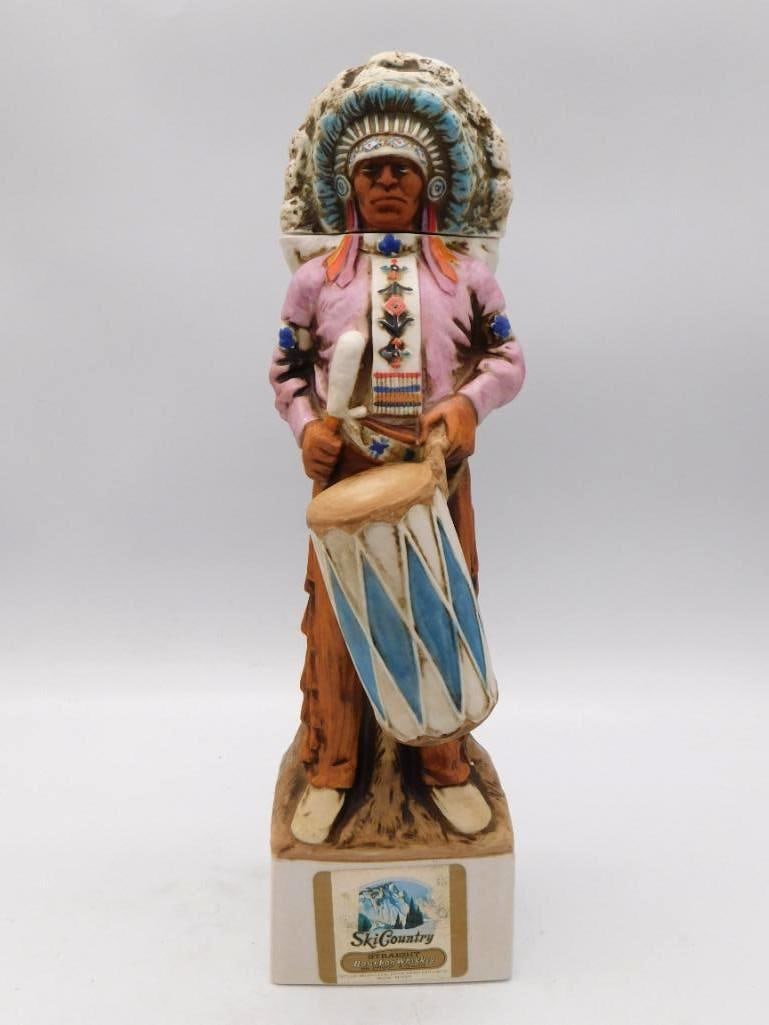 Ski Country Plains Indian Drummer: A handsome Ski Country decanter sculpted as a Plains Indian drummer in pink and orange buckskin, a hand drum bearing a blue diamond device held at his side and a feathered headdress upon him. Rises