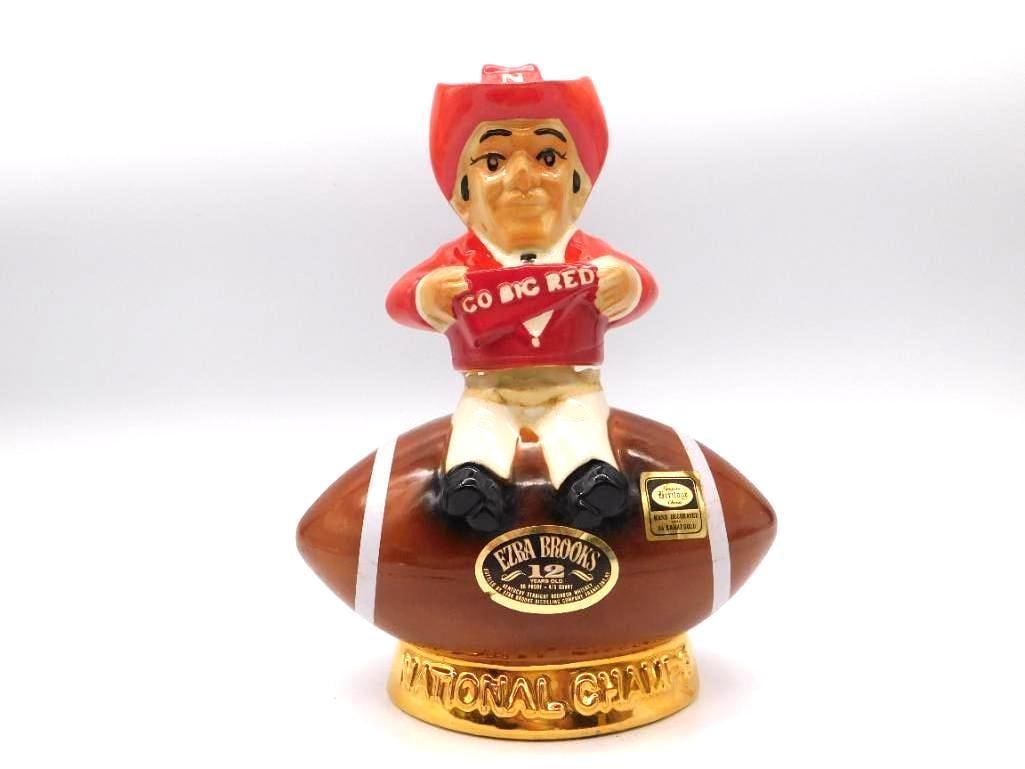 Ezra Brooks Nebraska Cornhuskers Go Big: A spirited Ezra Brooks decanter raised in tribute to Nebraska football, modeled as a Cornhusker mascot hoisting a Go Big Red banner atop a football. The gold-tone base carries the National Champions