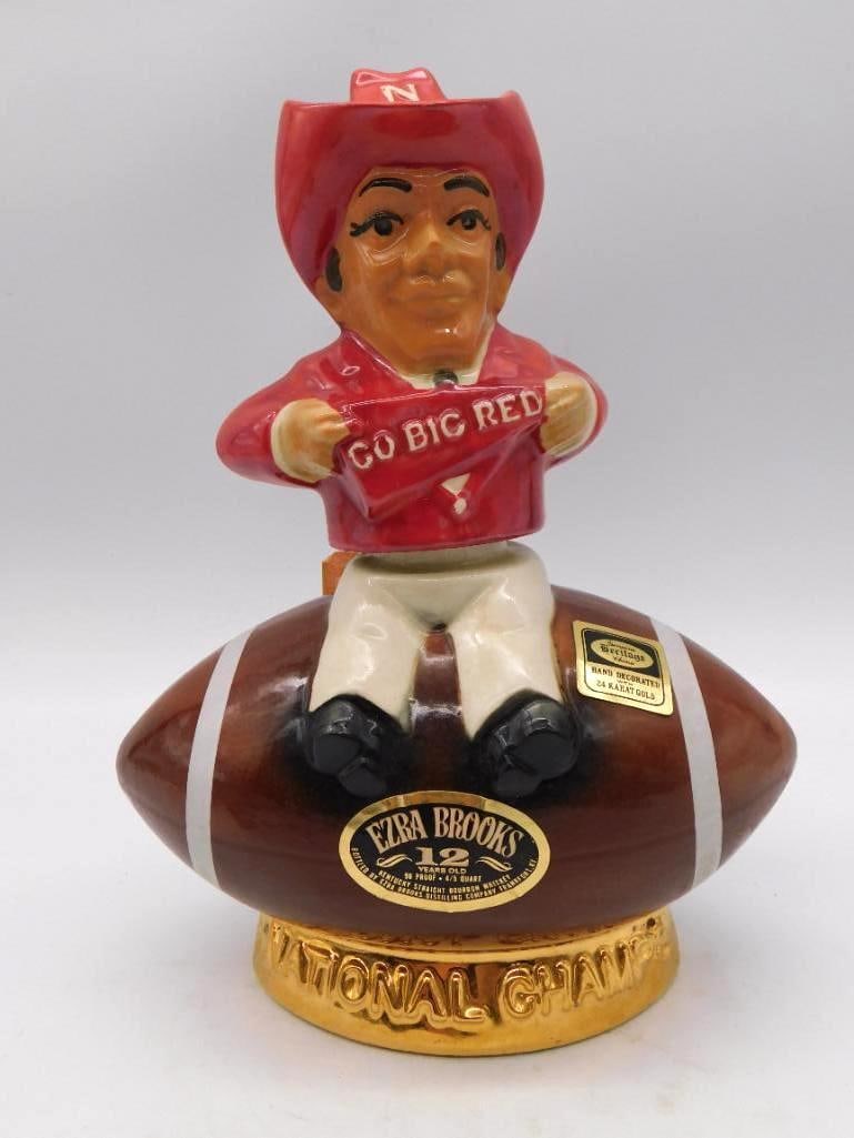 Ezra Brooks Go Big Red Nebraska Natl: An Ezra Brooks 12 Years Old decanter celebrating Nebraska football's National Championship, modeled as a red-hatted partisan hoisting a "Go Big Red" banner atop a football base. Hand-decorated with