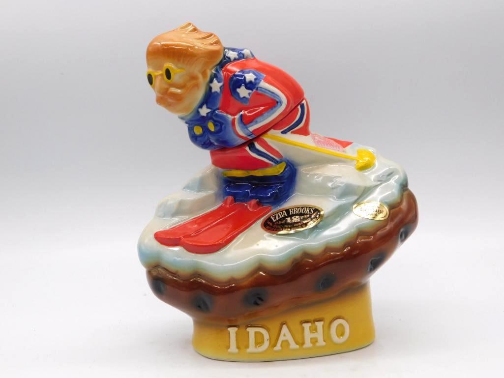 Ezra Brooks Heritage China Idaho Skier: An Ezra Brooks Heritage China decanter sculpted as a downhill skier in a red, white and blue suit, carving across a snowy mountain base marked Idaho. The sculptor caught the lean of a good run.