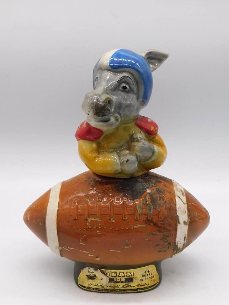 Jim Beam Football Pig Mascot Ceramic: A ceramic Jim Beam decanter modeled as a pig mascot in a blue and white football helmet and jersey, seated atop a football-shaped base labeled Beam 100 Kentucky Straight Bourbon Whiskey. Approximately