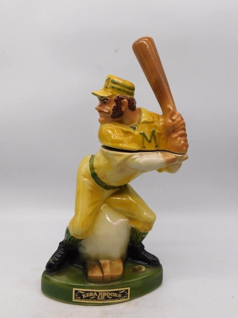 Ezra Brooks Baseball Batter Decanter in: A spirited Ezra Brooks decanter modeled as a ballplayer squared at the plate, bat cocked and eye on the pitcher, his mustache as stout as his swing. The uniform is painted yellow and green with the le