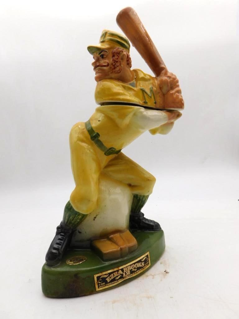 Ezra Brooks Baseball Batter 12 Years Old: An Ezra Brooks 12 Years Old decanter sculpted as a baseball batter of the old style, caught mid-swing in a yellow uniform bearing an M monogram, upon a green oval base. The sculptor got the swing