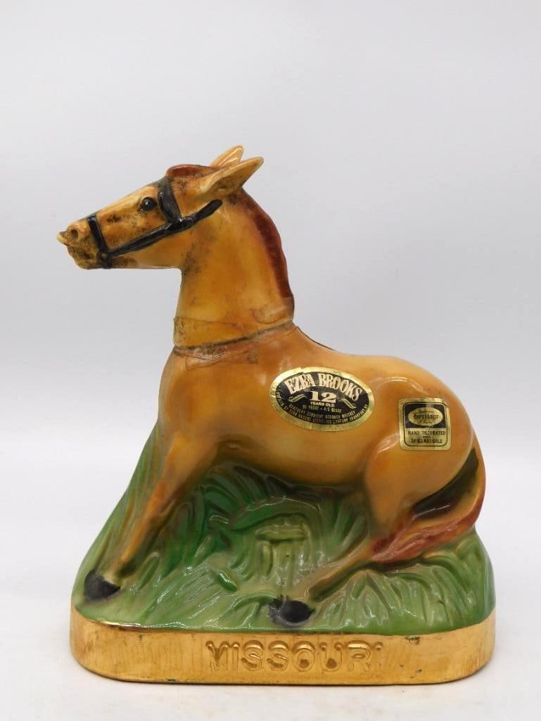 Ezra Brooks Missouri Mule Reclining: An Ezra Brooks decanter modeled as a Missouri mule at rest in the grass, bridled and alert of ear, the whole set upon a gold-tone plinth lettered Missouri. Hand decorated in the Heritage China manner