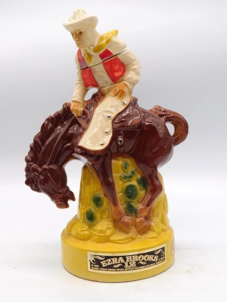 Ezra Brooks Pioneer Settler and Horse: An Ezra Brooks decanter sculpted as an early American pioneer or settler couple standing beside a horse's head, worked in cream and brown tones upon a textured gold base. The base carries the Ezra Bro