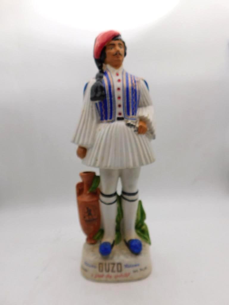 Metaxa Ouzo Greek Evzone Guard: A decanter modeled as a Greek Evzone guard in pleated white fustanella, blue embroidered vest and red cap, an amphora and laurel at his feet. The costume detail is handsome, though two of the