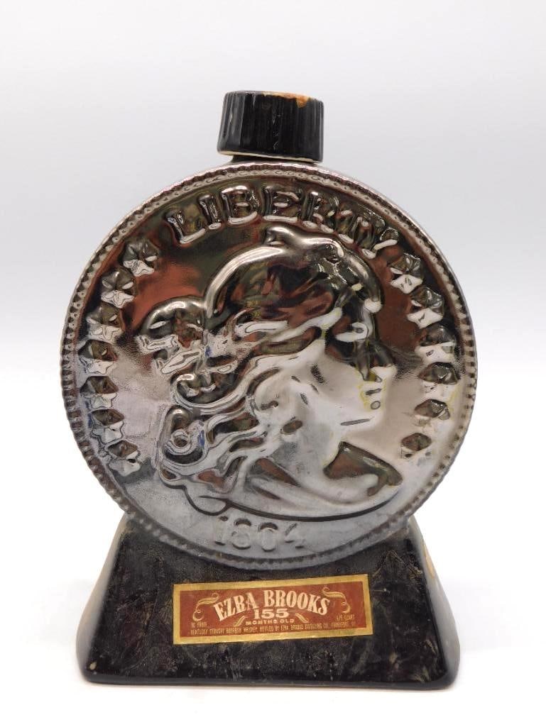 Ezra Brooks Liberty Coin Silver-Tone: An Ezra Brooks ceramic decanter molded as an oversized Liberty coin dated 1804, finished in silver-tone upon a black base. The gold paper label reads Ezra Brooks 155 Months Old Kentucky Straight Bourb
