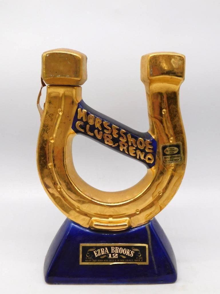 Ezra Brooks Horseshoe Club Reno 24 Karat: A jaunty Ezra Brooks decanter shaped as a lucky horseshoe in 24 karat gold over cobalt, the club name lettered boldly across the center bar. It stands upon a deep blue plinth bearing the Ezra Brooks