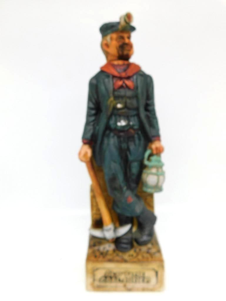 Old Time Coal Miner Decanter with Pick: An earnest decanter modeled as an old-time collier in cap and lamp, leaning on his pick with a lantern in his free hand and two crates of Permissible Collier's Dynamite stacked at his back. The