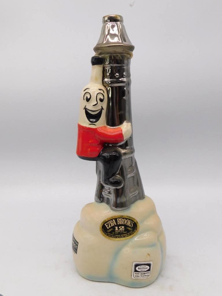 Ezra Brooks Platinum Oil Derrick: An uncommon Ezra Brooks decanter modeled as a genial bottle-headed fellow in a red jacket clinging to a platinum-luster oil derrick, the whole set upon a pale mound. The grin is frank and the luster