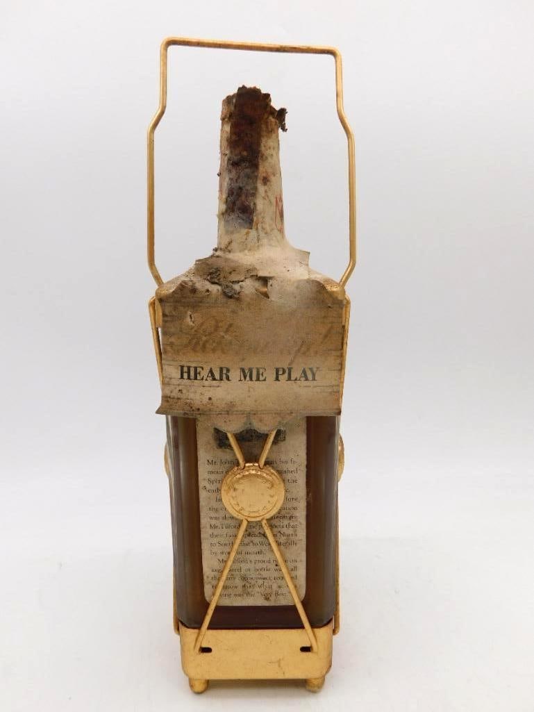 Unmarked Glass Decanter in Gilt Carrying: A square glass whiskey bottle held in a gilt wire cradle with swing handle and applied seals. The paper neck banner is printed Hear Me Play, while the body label is so effaced that the distiller's