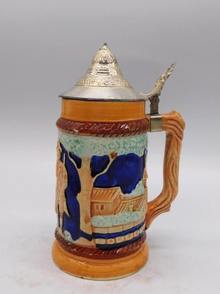 German-Style Bavarian Stein Ceramic: A ceramic whiskey decanter styled after a Bavarian beer stein, worked in cobalt blue relief with a village scene, fitted with a wood-grain handle and a hinged pewter lid. A sturdy and agreeable