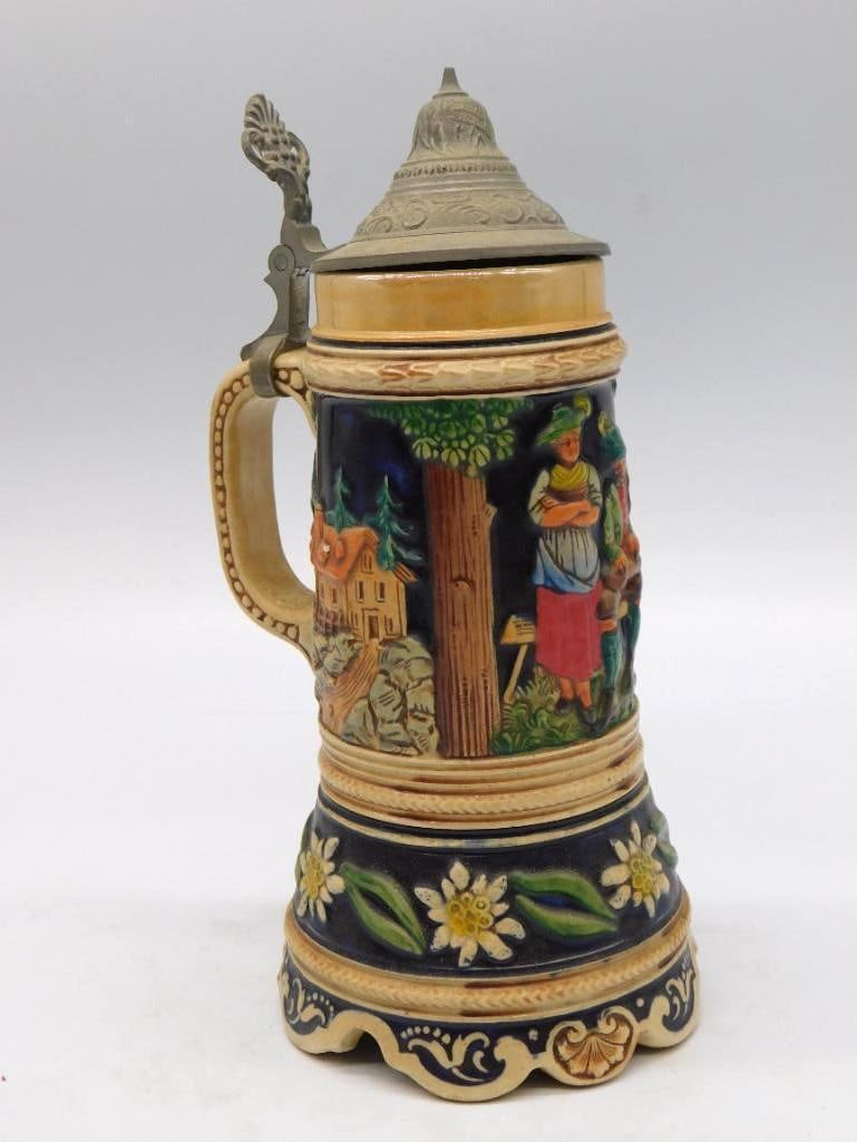German Bavarian Ceramic Beer Stein with: A vintage German-made ceramic beer stein with a hinged pewter lid, worked in relief with a Bavarian alpine village scene, figures in traditional dress, and edelweiss about the base. A stout and